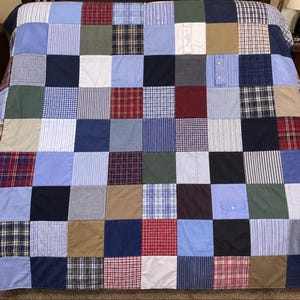 May include: A patchwork quilt constructed from square fabric panels featuring various patterns including stripes, plaids, and solid colors in shades of blue, red, white, green, brown, and black. Some squares include visible shirt buttons and pockets.