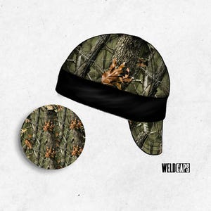 May include: A welding cap featuring a forest camouflage pattern with tree bark and brown leaf details, a solid black band, and a circular inset showing the fabric texture. The text WELD CAPS is printed in black in the bottom right corner.