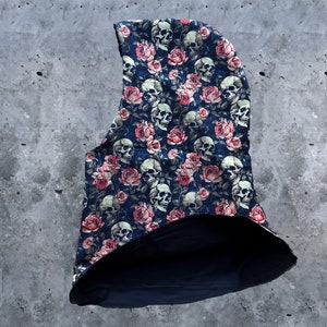 Pode incluir: Uma balaclava preta com capuz com uma estampa de caveiras e rosas. O tecido é azul escuro com caveiras brancas e rosas cor-de-rosa.