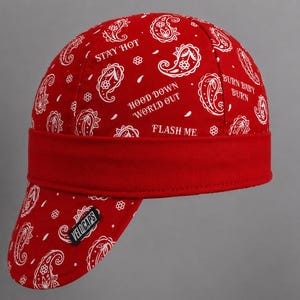 May include: A red welding cap featuring a white paisley pattern and the text STAY HOT, HOOD DOWN WORLD OUT, and BURN BABY BURN. The cap includes a solid red band and a black VELOCAPS label on the brim, providing a structured fit for head protection.