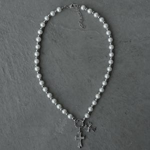 May include: A silver-toned necklace featuring a strand of white faux pearls and small silver beads. The necklace has two cross pendants, one larger and ornate, and a smaller, simpler cross charm.