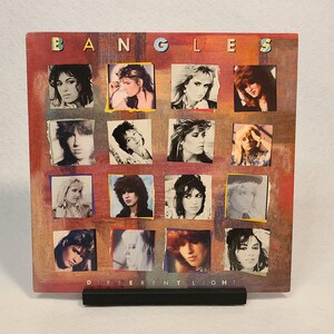 Puede incluir: Una portada de álbum de vinilo vintage con una banda llamada "The Bangles". La portada presenta un collage de fotos de los miembros de la banda, con el título "Different Light" impreso en la parte inferior.