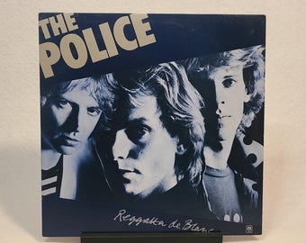May include: A blue vinyl record album cover featuring a band called "The Police". The album title is "Reggatta de Blanc". The cover features three men with their faces partially obscured by a diagonal blue line.