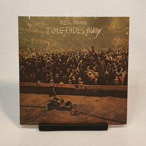 Puede incluir: Una portada de álbum de vinilo vintage con Neil Young. La portada muestra una multitud de personas en un concierto con el título "Time Fades Away" en letras blancas.