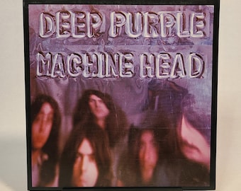 May include: A framed album cover for Deep Purple's "Machine Head." The album art features the band members in a blurred image with the album title and band name in a raised, white font against a purple background. The frame and stand are black.