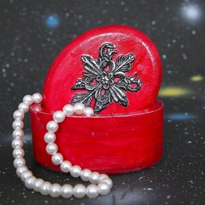 May include: A red oval-shaped jewelry box with a silver floral design on the lid. A strand of white pearls is draped around the box.