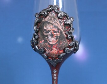 May include: A red wine glass with a skull design in a silver frame. The glass has a silver glitter finish and rhinestones on the stem.