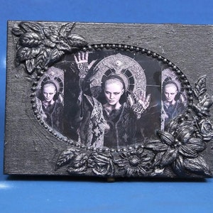 May include: A rectangular, gray decorative box with a black and white image of a person with their hands raised. The image is framed with an oval border and surrounded by silver floral embellishments.