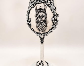 May include: A decorative wine glass with a black and white illustration of a horned figure. The glass features a detailed, ornate frame around the image and a red gem at the base. The stem and base are white.
