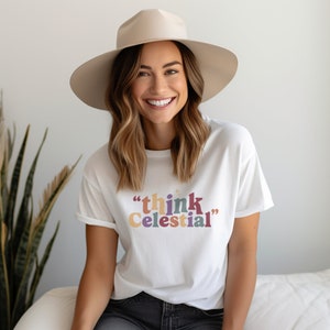 May include: White t-shirt with the text "think Celestial" in colorful letters. The shirt is worn by a person with long brown hair and a beige hat.