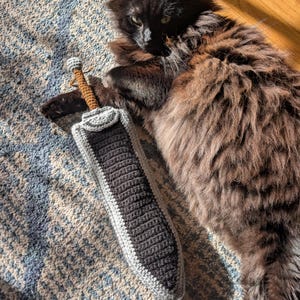 May include: A crocheted toy sword featuring a dark charcoal gray blade with a light gray border. The handle is brown with a small light gray pommel at the end. The sword is crafted with a textured yarn stitch pattern and has a rounded tip.