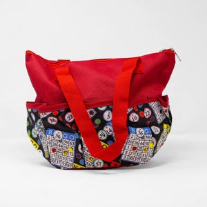 Peut inclure: Un sac fourre-tout rouge avec une section inférieure noire ornée d'un motif de boules et de cartons de bingo colorés. Le sac possède deux anses en tissu rouge, une fermeture éclair sur le dessus et des poches latérales. Les cartons affichent BINGO.