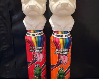 May include: Two novelty dog-shaped items. The white dog heads sit atop colorful cylindrical bases. One base is red with the text "BE A WINNER! SOYEZ GAGNANT!" and a pink elephant graphic. The other is orange with the same text and graphics.