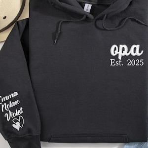 Peut inclure: Sweat-shirt à capuche noir avec du texte blanc qui dit "opa Est. 2025" sur le devant et du texte blanc qui dit "Emma Nolan Violet" avec deux cœurs sur la manche gauche.