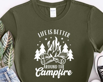 May include: Olive green t-shirt with the text "LIFE IS BETTER AROUND THE CAMPFIRE" in white. The graphic features a campfire with flames, logs, and trees. The shirt is paired with blue jeans and black canvas sneakers.