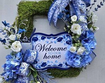 May include: A blue and white floral wreath with a sign that says "Welcome to our home". The wreath is decorated with blue and white ribbon, white roses, and blue hydrangeas.