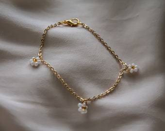 May include: A gold-colored chain bracelet with three small daisy flower charms. Each daisy has white petals and a gold center. The bracelet is displayed on a cream-colored, textured surface.