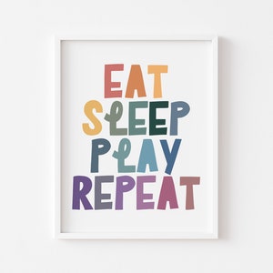 Könnte beinhalten: Ein weißer gerahmter Druck mit dem Text "Eat Sleep Play Repeat" in bunten, stilisierten Buchstaben.