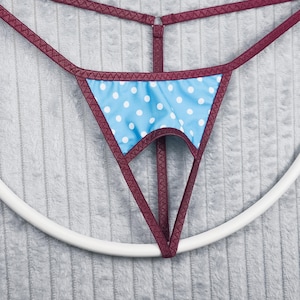 Op de afbeelding: Een string met een lichtblauw voorpaneel met witte stippen, omlijst door een bordeauxrode rand. De bandjes en randen zijn bordeauxrood. De string wordt opgehangen door een wit cirkelvormig object.