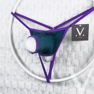 May include: A teal and purple thong-style garment with a white, rounded insert. The thong is suspended within a white circular frame. A small tag with a stylized "M" logo is attached.