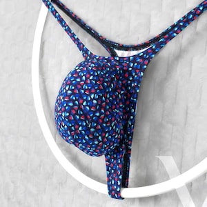 May include: A thong featuring a contoured pouch with a dark blue base color and a repeating pattern of small, irregular red and light blue shapes. The garment includes thin, matching fabric straps that extend from the pouch to form the waistband.