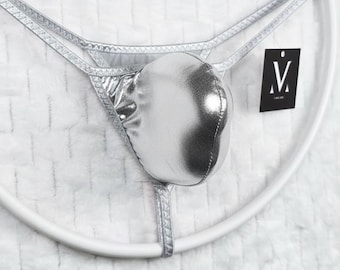 May include: A metallic silver thong featuring a contoured front pouch and thin, textured side straps. A small black square tag with the white letter M and a dot above it is attached to the side. The garment is displayed on a white circular frame.