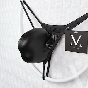May include: Black thong underwear with a pouch. The underwear is made of a shiny material and features a tag with a white "V" logo on a black background. The underwear is suspended on a white circular frame.