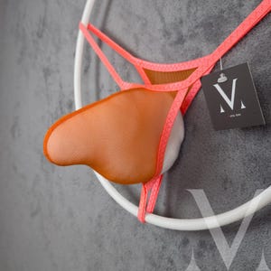 Op de afbeelding: Een neonoranje en roze string-ondergoed met een mesh zakje en een witte binnenkant. Het kledingstuk is opgehangen aan een wit cirkelvormig frame. Een label met een logo is bevestigd.