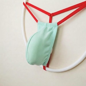 Op de afbeelding: Een mintgroene string met een buidelvormig voorpand en dunne rode elastische bandjes. Het kledingstuk heeft een centraal gevormd stoffen voorpand in een lichtgroene tint, verbonden met een rode tailleband en zijbandjes.