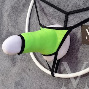 May include: A novelty item featuring a plush, white phallus with a neon green cover and black trim. The item is suspended by a white ring and black straps, with a tag visible in the background. The design is suggestive and playful.