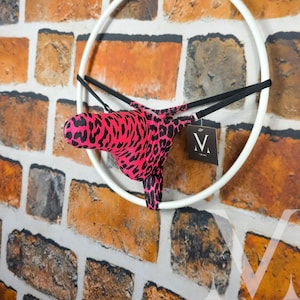 Op de afbeelding: Een nieuwigheid met een roze en zwarte luipaardprint, opgehangen in een witte cirkelvormige lijst. Zwarte bandjes bevestigen het item. Een label met de letter "V" is bevestigd. De achtergrond is een bakstenen muur.
