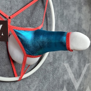 May include: A teal and red lingerie item with a white padded insert. The item features a metallic teal fabric with red trim and straps. A white circular support structure is visible, along with a tag.