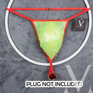 May include: A lime green thong with red trim and a red string with a red jewel at the end. The thong is hanging on a white ring. The text "PLUG NOT INCLUDED" is printed below the ring.