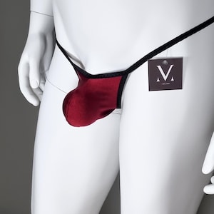 May include: A burgundy thong with black trim and a black string waistband. A small tag with a white "V" logo hangs from the waistband. The thong is displayed on a white mannequin.