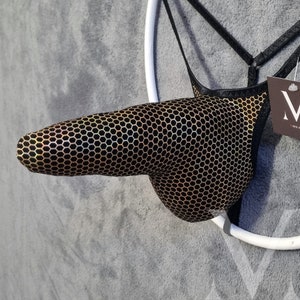 May include: A black and gold patterned thong with a honeycomb design. The thong is suspended from a white circular frame and black straps. A tag with the letter "M" is attached to the garment.