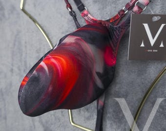 Op de afbeelding: Een close-up van een zwembroek met een wervelend patroon in rood, zwart en wit. Het kledingstuk heeft dunne bandjes en een label met de tekst "mens lew". De zwembroek wordt gepresenteerd op een goudkleurige hanger tegen een grijze achtergrond.