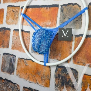 Può includere: Perizoma blu con stampa leopardata, esposto su una cornice circolare bianca. La biancheria intima è realizzata in un tessuto trasparente e ha spalline blu. È visibile una piccola etichetta con la lettera "V".