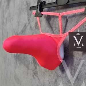 May include: Bright pink underwear with a unique design, hanging from a black hanger. The garment has a distinctive shape and a contrasting white interior. A tag with the letters "V" and the text "mens lew" is attached.
