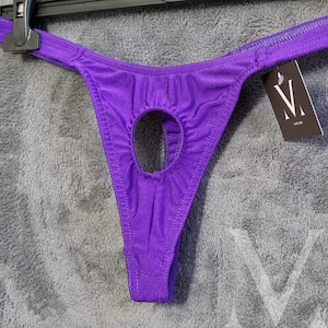 Könnte beinhalten: Nahaufnahme eines lilafarbenen Tangas mit Schlüsselloch-Design. Der Tanga ist aus glänzendem Stoff gefertigt und hängt an einem schwarzen Kleiderbügel. Ein Etikett mit weißem "V"-Logo ist angebracht.