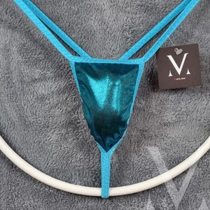 Può includere: Slip in tanga di colore blu turchese metallizzato con bordino turchese. La biancheria intima ha un piccolo cartellino attaccato con la lettera "M" e le parole "mens sew".