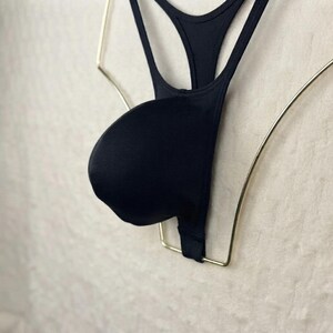 May include: A solid black thong with a contoured front pouch design, featuring a minimalist construction with thin straps that extend upward and downward, supported by a thin gold-toned metal frame structure for display purposes.
