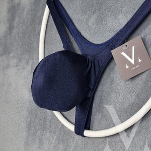 Könnte beinhalten: Ein marineblauer String-Unterwäsche mit Beutel und dünnen Trägern. Das Kleidungsstück hängt an einem weißen kreisförmigen Kleiderbügel. Ein kleines Etikett mit dem Text "mens-lew" ist angebracht.