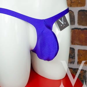 Pode incluir: Um close-up de uma tanga roxa vibrante. A roupa íntima é exibida em um manequim branco. A tanga tem uma bolsa na frente e uma cintura fina. Uma pequena etiqueta com um logotipo V branco está anexada.