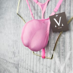 May include: A pink lingerie set with a sheer, rounded front and thin straps. The set is displayed on a gold-coloured metal hanger. A tag with a stylized "M" logo hangs from the strap.