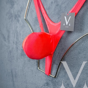 May include: A bright pink,  swimsuit with a V-shaped front and a single strap on each side. The swimsuit has a padded cup on the front. The swimsuit is hanging on a gold hanger with a black tag that says "V by me".