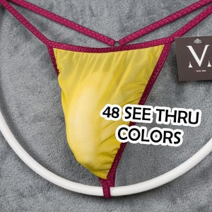 May include: Yellow see-through thong with burgundy trim and straps. The text "48 SEE THRU COLORS" is displayed on the front. A white circular object is in the foreground, and a tag is in the background.