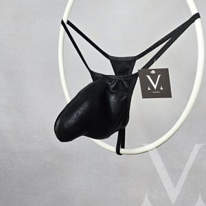 Op de afbeelding: Zwarte string met een metallic glans, opgehangen aan een wit cirkelvormig frame. Het kledingstuk heeft een zakachtige voorkant en dunne bandjes. Een label met een gestileerd "M"-logo hangt aan het frame.