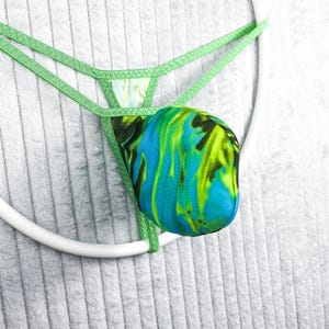 Peut inclure: Un string vert avec un motif abstrait vibrant dans les tons de bleu, vert et noir. Le string est suspendu à une structure circulaire blanche sur un fond gris clair texturé. Le design présente une impression unique et accrocheuse.