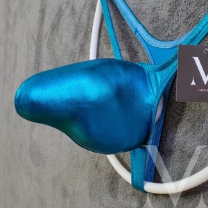 Könnte beinhalten: Ein türkisfarbener, metallischer String-Badeanzug mit weißem Ringdetail. Der Badeanzug hat eine taschenartige Vorderseite und dünne Träger. Ein schwarzes Etikett mit weißem "M"-Logo ist angebracht.