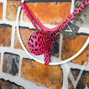 Op de afbeelding: Een roze en zwarte string badpak met luipaardprint hangend aan een witte cirkelvormige hanger. Het badpak is gemaakt van een rekbaar materiaal en heeft een levendige roze basiskleur met zwarte vlekken. Een label met de letter "V" is bevestigd.